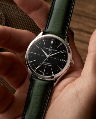 Maennerhand haelt eine Clifton Baumatic Uhr von Baume & Mercier in Stahl mit schwarzem Zifferblatt und gruenem Lederarmband in der Handflaeche