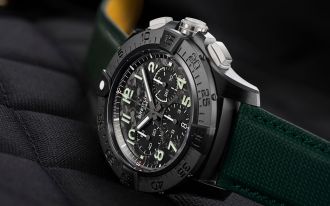 Breitling Avenger Night Mission Uhr mit schwarzem Keramikgehaeuse, schwarzem Camouflage-Zifferblatt und dunkelgruenem Textilarmband seitlich auf schwarz gesteppten Stoffhintergrund liegend