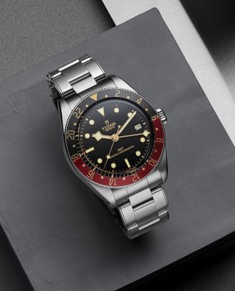 TUDOR Black Bay GMT in Stahl mit schwarz-roter Luenette und schwarzem Zifferblatt auf grauem Hintergrund liegend