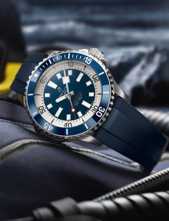 Breitling Superocean Uhr in Stahl mit blauer Luenette, blauem Zifferblatt und blauem Kautschukarmband seitlich liegend auf Lifestylehintergrund