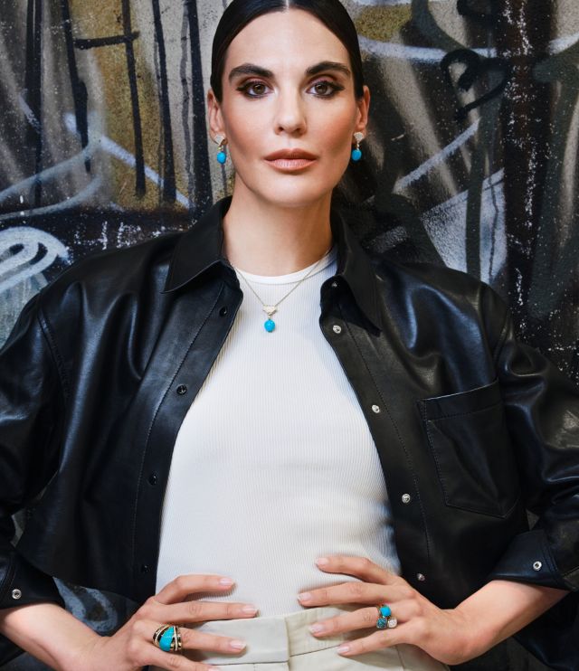 Frau mit türkisfarbenem Schmuck aus der Chromatic-Kollektion des Atelier Mühlbacher, kombiniert mit Lederjacke und schlichtem Oberteil vor künstlerischem Hintergrund.