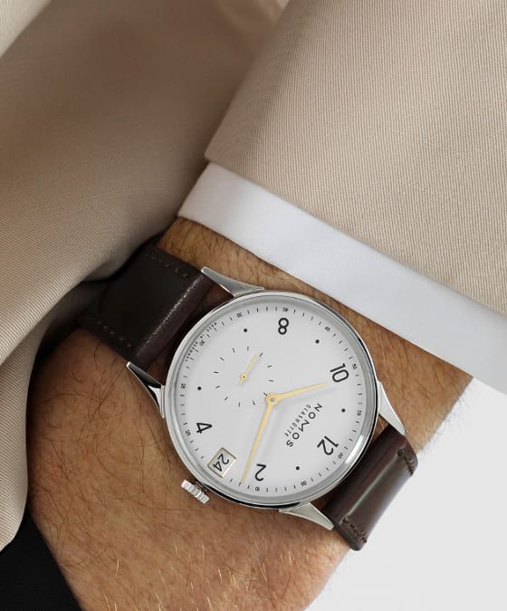 Herrenhandgelenk mit einer Minimatik Uhr von NOMOS Glashuette in Stahl mit weissem Zifferblatt, goldenen Applikationen und einem braunem Lederarmband