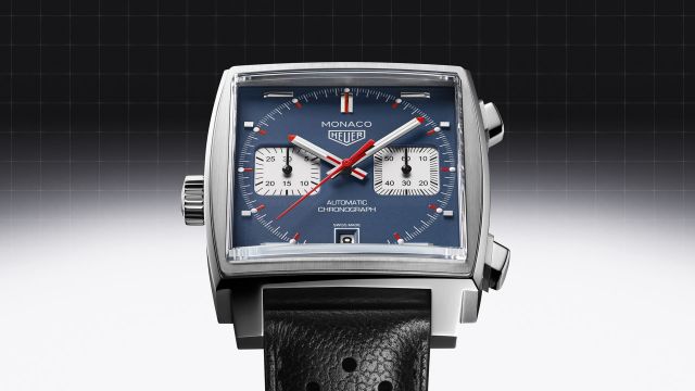 TAG Heuer Monaco Chronograph mit Stahlgehaeuse, blauem Zifferblatt und scwharzem Lederarmband vor schwarz-grauem Hintergrund