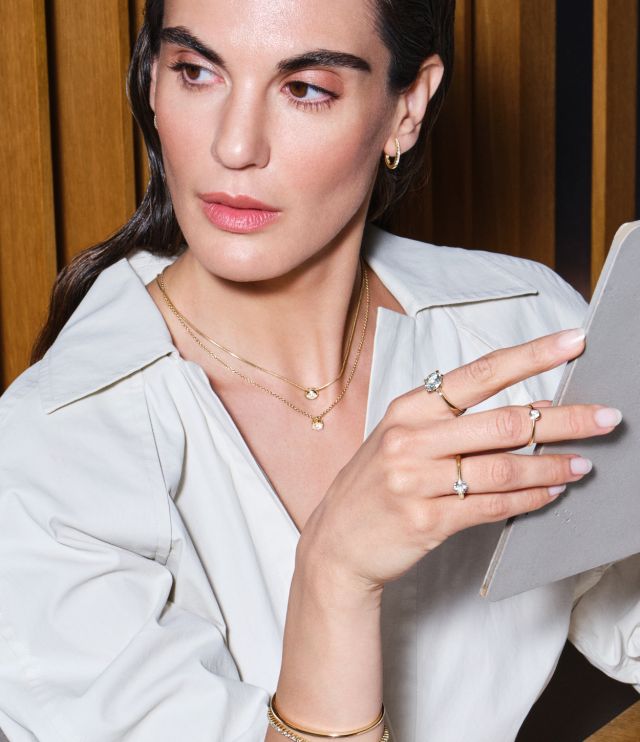 Frau mit Goldschmuck aus der One-Kollektion des Atelier Mühlbacher: zarte Ringe, zwei feine Halsketten und ein elegantes Armreif-Design, stilvoll in Szene gesetzt.