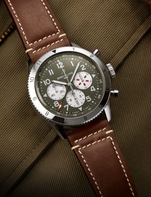 Breitling Classic AVI Chronograph in Stahl mit militarygruenem Zifferblatt und braunem Lederarmband auf khakifarbenem Textilhintergrund liegend