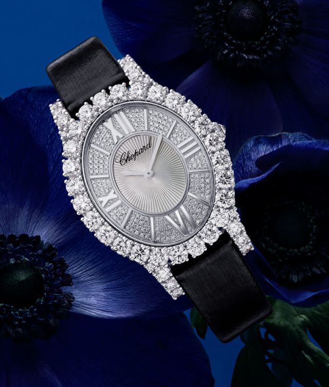 Chopard L'Heure du Diamant Damenuhr mit Gehaeuse in Weissgold besetzt mit Diamanten, silbernem Zifferblatt mit roemischen Ziffern und schwarzem Textilarmband vor blauem Hintergrund