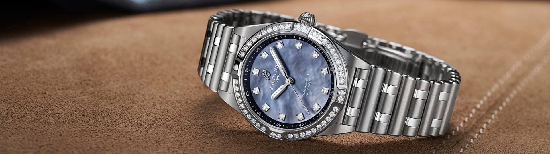 Breitling Chronomat 28 Uhr in Stahl mit Diamantluenette, Rouleauxarmband und Zifferblatt aus blauem Perlmutt seitlich auf braunem Textiluntergrund liegend