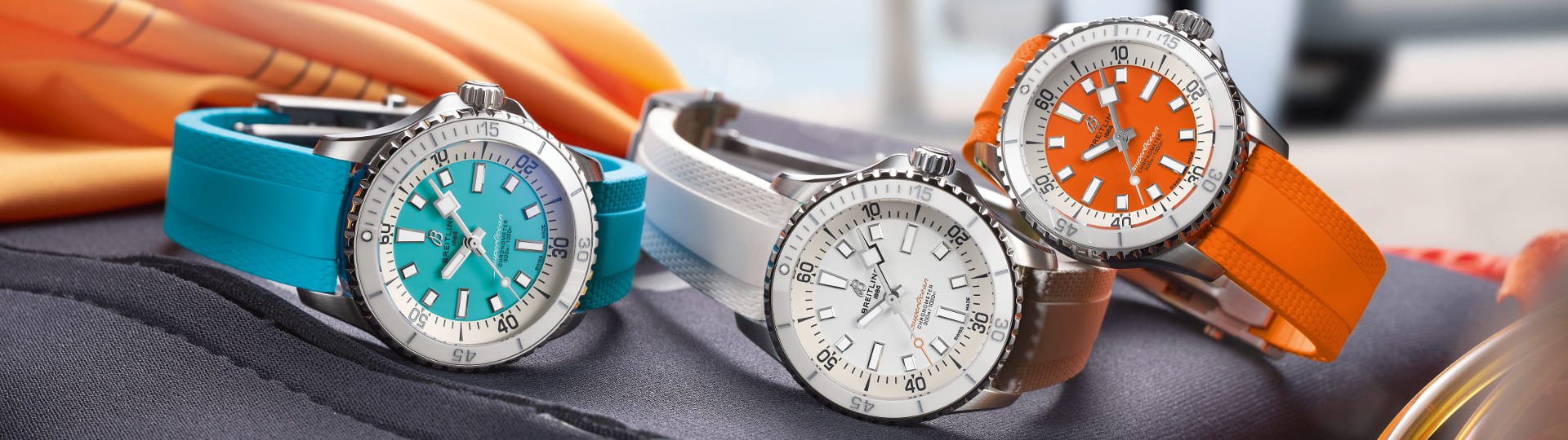 Drei Breitling Superocean Uhren mit Stahlgehaeusen, Zifferblaetter in tuerkis, weiss und orange und farbig identischen Kautschukarmbaender nebeneinander auf Lifestylehintergrund liegend