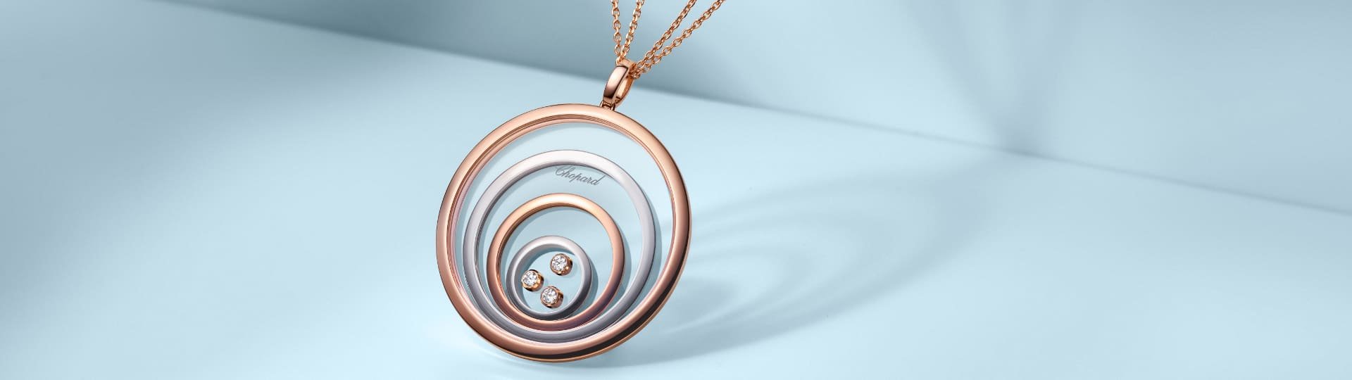 Chopard Happy Spirit Anhaenger aus mehreren Kreisen in Rose- und Weissgold, im innersten Kreis drei bewegliche Diamanten, an einer feinen zweireihigen Rosegoldkette vor hellblauem Hintergrund