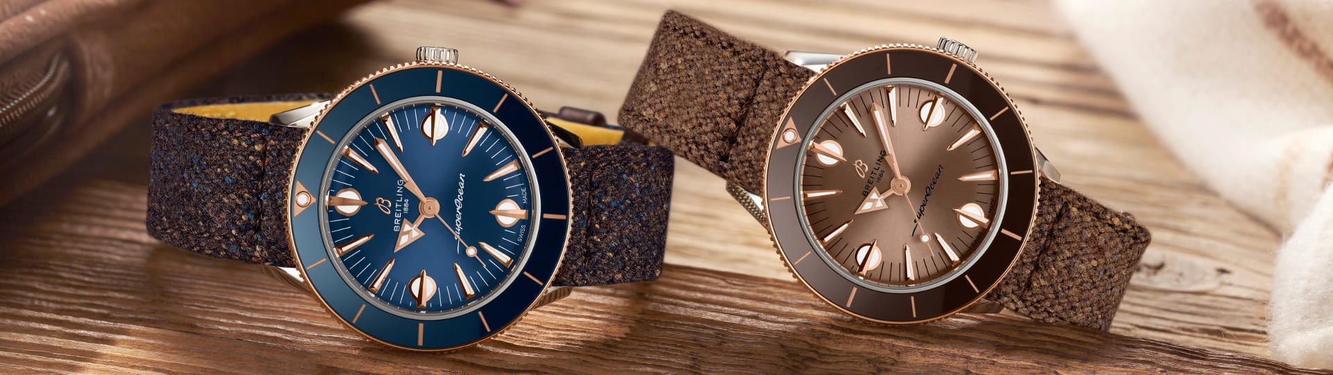 Zwei Superocean Heritage Uhren von Breitling in Stahl und Rotgold mit blauem und braunem Zifferblatt und Luenette und Textilbaendern liegend auf einem Holzuntergrund