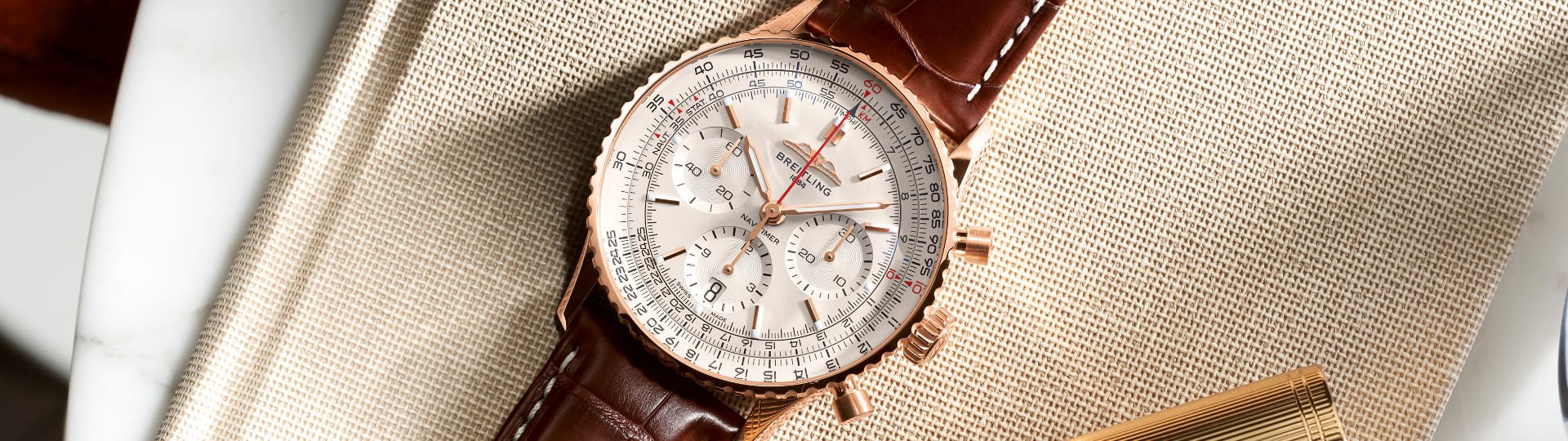 Breitling Navitimer Chronograph in Gold mit cremefarbenem Zifferblatt und braunem Lederarmband auf beigefarbenem Stoff liegend