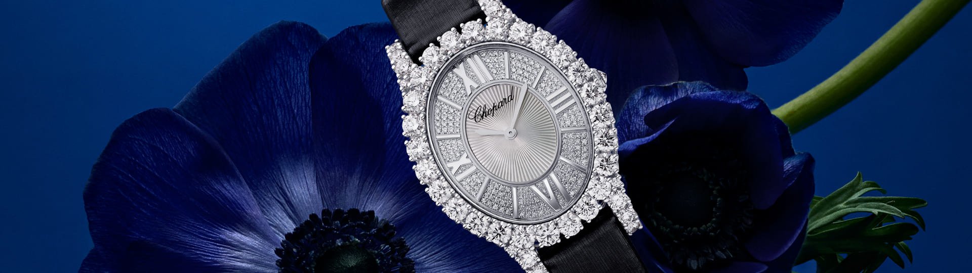 Chopard L'Heure du Diamant Damenuhr mit Gehaeuse in Weissgold besetzt mit Diamanten, silbernem Zifferblatt mit roemischen Ziffern und schwarzem Textilarmband vor blauem Hintergrund