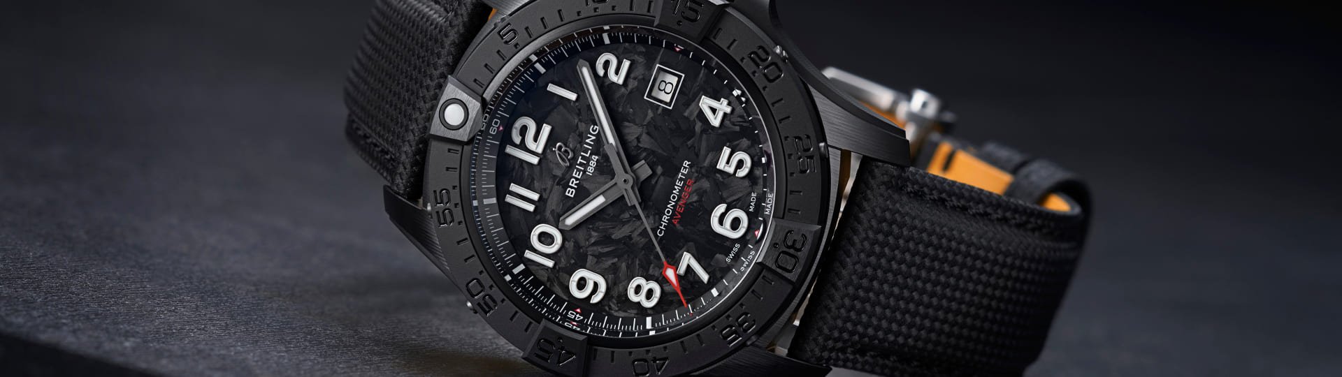 Breitling Avenger Night Mission Uhr mit schwarzem Gehaeuse, Zifferblatt und Textilarmband seitlich auf dunklem Untergrund liegend