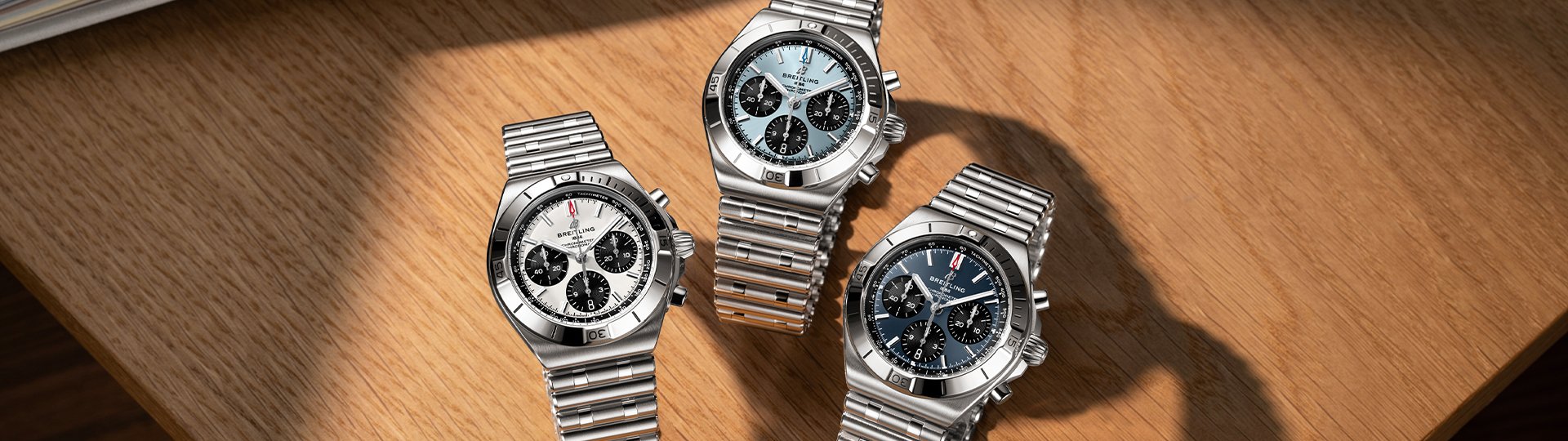 Drei Breitling Chronomat B01 42 Chronographen mit weißen, eisblauen und dunkelblauen Zifferblättern auf heller Holzoberfläche.