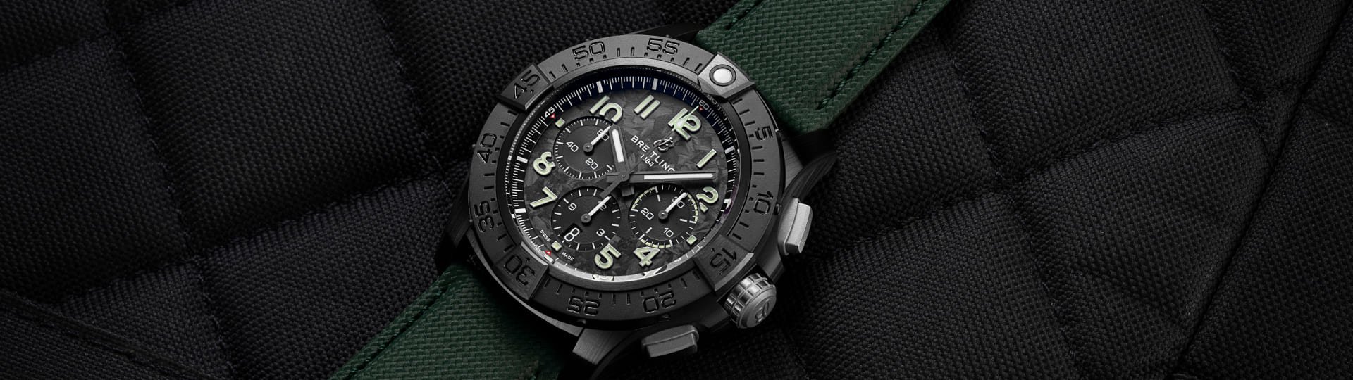 Breitling Avenger Chronograph Night Mission mit scharzem Keramikgehaeuse, schwarzem Zifferblatt und dunkelgruenem Textilband auf schwarzem gesteppten Stoffhintergrund liegend