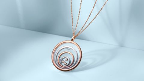 Chopard Happy Spirit Anhaenger aus mehreren Kreisen in Rose- und Weissgold, im innersten Kreis drei bewegliche Diamanten, an einer feinen zweireihigen Rosegoldkette vor hellblauem Hintergrund