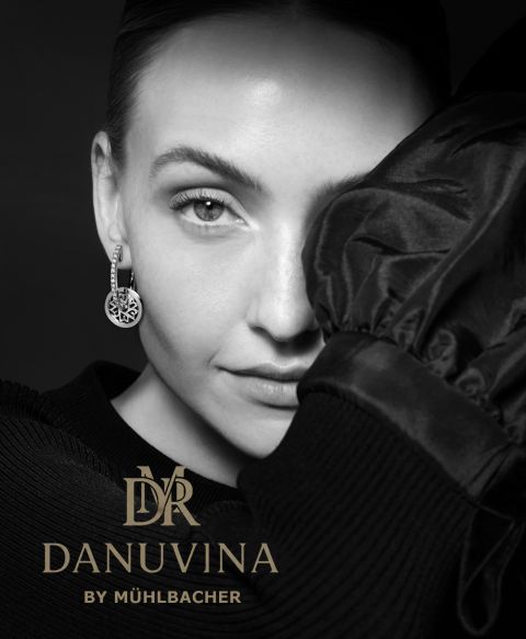 Nahaufnahme einer Frau mit edlen Danuvina Ohrringen by Mühlbacher, schwarz-weiß fotografiert, kombiniert mit dem goldfarbenen Markenlogo vor dunklem Hintergrund. Nahaufnahme einer Frau mit edlen Danuvina Ohrringen by Mühlbacher, schwarz-weiß fotografiert, kombiniert mit dem goldfarbenen Markenlogo vor dunklem Hintergrund.