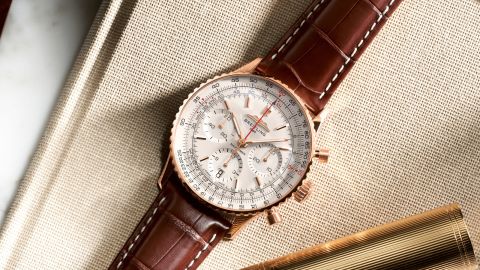 Breitling Navitimer Chronograph in Gold mit cremefarbenem Zifferblatt und braunem Lederarmband auf beigefarbenem Stoff liegend Breitling Navitimer Chronograph in Gold mit cremefarbenem Zifferblatt und braunem Lederarmband auf beigefarbenem Stoff liegend