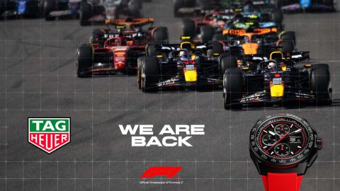 Tag Heuer Formula 1 Uhr mit schwarzem Gehaeuse und rotem Kautschukband vor Formula 1 Rennautos und links im Bild Tag Heuer Logo mit We are back Schriftzug