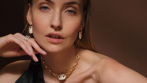 Nahaufnahme einer Frau mit goldener Crivelli-Halskette mit pavébesetztem Anhänger, passenden Ohrringen und diamantbesetzten Ringen an der linken Hand. Nahaufnahme einer Frau mit goldener Crivelli-Halskette mit pavébesetztem Anhänger, passenden Ohrringen und diamantbesetzten Ringen an der linken Hand.