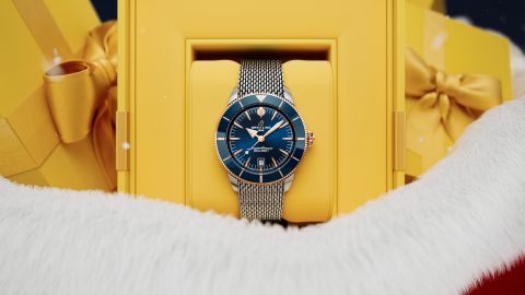 Nahaufnahme einer blauen Breitling Superocean Uhr in einer gelben Geschenkbox, eingerahmt von weißem Winterfell und goldenen Geschenkpaketen im Hintergrund. Nahaufnahme einer blauen Breitling Superocean Uhr in einer gelben Geschenkbox, eingerahmt von weißem Winterfell und goldenen Geschenkpaketen im Hintergrund.